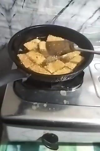 A close-up shot shows a black frying pan filled with numerous rectangular pieces of food, likely fried tofu, submerged in hot oil. The food is golden brown and appears to be sizzling. A wooden spatula is actively stirring the pieces in the pan. The pan sits on a silver gas stove with a black burner. The background features a marbled backsplash. The video is shot from a slightly elevated angle.