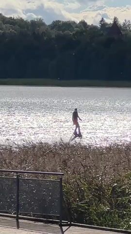 A person is wakeboarding on a lake. The sun is shining and reflecting off the water, creating a shimmering effect. There are trees and a building in the background. The person is wearing a dark-colored swimsuit and is being propelled by a device attached to their feet. The device is spraying water, creating a wake behind them.