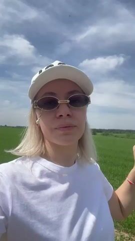 A blonde woman wearing a white "ICON" baseball cap, white t-shirt, and oval sunglasses with a pearl dangle earring on her right ear stands in a green field. She looks at the camera, raises her left hand, then lowers it to touch her cap and the back of her head. The background shows a vast green field under a cloudy sky.