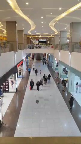 A wide shot captures a mall interior with a long, open walkway. The ceiling features illuminated, wavy panels. Several shops are visible on either side of the walkway, with their storefronts displaying merchandise and signage. A few people are walking down the walkway in various directions. On the left, a shop with a display of shoes is visible. On the right, there are several more shops, including one with a sign that appears to say "McDonald's". The floor of the walkway is tiled in a light color. The overall impression is of a typical shopping mall environment.