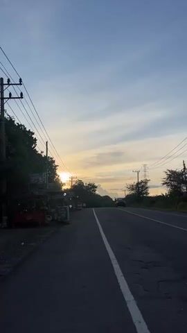The video shows a paved road with a white line down the center, bordered by trees and power lines. A person on a scooter is riding away from the camera down the road. The sun is setting, casting a warm glow across the sky, which is a pale blue with streaks of orange and yellow clouds. There are small roadside stalls on the left side of the road. A few other vehicles are visible in the distance. The overall atmosphere is peaceful and serene.