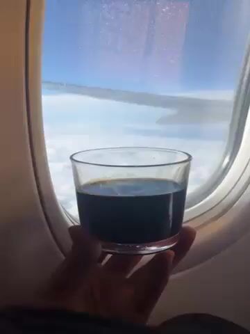 A hand holds a glass of black liquid in front of an airplane window. Beyond the glass and window, part of the wing of the airplane is visible, and beyond that is a blue sky dotted with clouds. The hand is pale-skinned. A child's voice can be heard.
