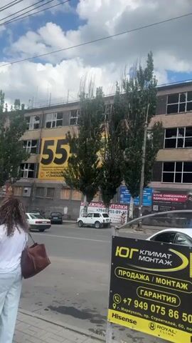 This video captures a street view in Mariupol, Ukraine. The main subject is a multi-story building with a facade of light brown tiles. Many of its windows are broken, suggesting damage from conflict. The building is adorned with various advertisements and signs.

In the foreground, a bright yellow and black sign promotes "ЮГ КЛИМАТ МОНТАЖ" (South Climate Installation) and "КОНДИЦИОНЕР" (Air Conditioner), offering sales, installation, and maintenance services. It displays phone numbers and an address in Mariupol. A white car is parked in front of this sign, with other cars visible on the street.

Behind the yellow sign, two tall, slender trees with green foliage stand in front of the damaged building. Several other trees are also present. The building itself features large advertisements, including one with the number "55" prominently displayed. Other signs visible are for "iPhone," "Samsung," and "TCL." Another sign advertises "ПАССАЖИРСКИЕ ПЕРЕВОЗКИ В ГРУЗИЮ" (Passenger transport to Georgia). Further signs include "ОБОИ КАРНИЗЫ" (Wallpaper Cornices) and "КРУГЛОСУТОЧНО" (24 Hours).

The sky is partly cloudy, indicating daytime. The overall scene suggests a street in a city that has experienced significant damage, with commercial activity continuing amidst the destruction. No people are clearly visible or identifiable in the video. The presence of damaged windows and ongoing commercial advertisements creates a stark contrast, hinting at the resilience and ongoing life in the city despite the hardships.