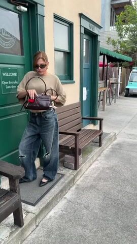 Here's a brief description of the video:

The video starts with a woman standing outside a building with green doors and beige walls, posing for a photo with a purse. The camera pans to the right to show a man standing on the sidewalk across the street, taking a photo of her with his phone. The camera then pans back to the woman, who resumes posing. The building has the address "506" on the door and a sign that reads "Alta Coffee & Roasting Co." The area appears to be a street with other buildings and cars in the background.  A small electric vehicle is parked nearby, along with some wooden carts.