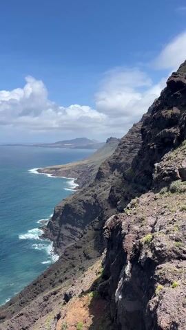 The video shows a scenic view of a coastline with rocky cliffs meeting the ocean. The sky is mostly blue with scattered white clouds. The water is a vibrant turquoise color with whitecaps visible where the waves crash against the shore. The camera pans from the rocky landscape on the right side to the open ocean in the center and back to a closer rocky cliff on the left. There's a brief shot showing a glass railing. The weather appears sunny with moderate winds.