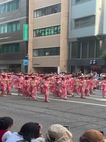 A large group of people in traditional Japanese attire, specifically pink kimonos with floral patterns and straw hats, march in a parade down a city street. They are holding what appear to be short, thin poles or batons. The street is lined with multi-story buildings, some with storefronts and signs in Japanese. Spectators are visible in the foreground, with the backs of their heads indicating they are seated or standing along the parade route. The weather appears to be sunny.