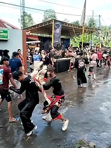 The video shows an outdoor performance with several people in traditional costumes. Two performers in the foreground are engaged in a dynamic dance, one wearing a black outfit and the other a costume with white and red elements that resembles an animal head. Behind them, a crowd of spectators watches the performance. There is a stage set up in the background with sound equipment and banners. The ground appears wet, possibly from rain or recent cleaning. The overall atmosphere suggests a cultural festival or performance event.