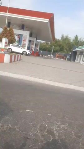 The video pans across a street view, showing a gas station with the "PERTAMINA" logo prominently displayed. Several cars and motorcycles are visible at the gas station. Adjacent to the gas station, a small convenience store with an orange and yellow awning is seen. Further down the street, a small yellow house with an Indonesian flag hanging vertically is visible. The sky is mostly clear with some clouds. The overall scene appears to be a typical street in an urban or suburban area.