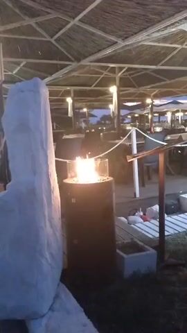 The video shows a dimly lit outdoor restaurant or bar area with a thatched roof. In the foreground, a modern gas fire pit with a glass enclosure burns, casting a warm glow. To the left, a large, pale object, possibly a statue or a covered piece of furniture, is visible. The background reveals rows of empty tables and chairs, suggesting the establishment is either closed or about to open for the evening. String lights are hung across the space, and a large, white air conditioning unit stands near some chairs. A sign above reads "BAR Veranda RESTAURANT." The overall atmosphere is relaxed and inviting, hinting at an evening dining or social experience.