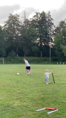 A young male is kicking a soccer ball on a grassy field. A toddler is standing to the right of the male, holding a pump. The male is wearing a white t-shirt and black shorts, and the toddler is wearing a light blue shirt and shorts. There are trees and a soccer goal in the background. The sky is cloudy.