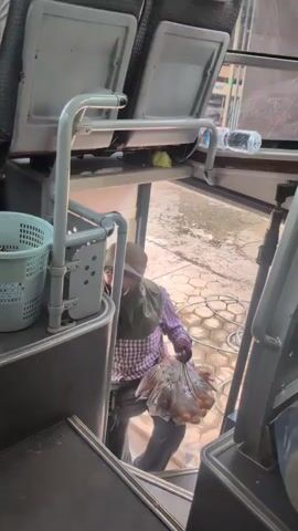This short video captures a candid moment inside a bus, likely in Pakxan, Laos. The primary subject is a person, dressed in a plaid shirt, a head covering, and gloves, who appears to be a vendor. They are holding several plastic bags filled with what look like small, round food items, possibly snacks or candies. The vendor is either boarding or disembarking the bus, stepping up from the outside into the aisle, near the entrance.

The setting is the interior of a bus, with rows of seats visible in the background. One seat in the foreground is empty, and another shows a person's shoulder and arm. A light-colored plastic basket is attached to a railing near the entrance, containing some small items. The floor outside the bus shows a patterned paving surface, and a metal railing runs along the side of the bus steps. A water bottle is visible on a shelf above the seats. The lighting suggests it could be daytime, possibly morning or afternoon, with natural light coming from the bus entrance and windows. The vendor's expression is a little ambiguous, but they appear to be focused on their task. The overall impression is one of everyday life and commerce on public transport in Laos.