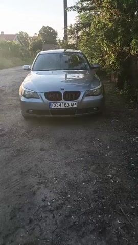 The video begins with a frontal view of a silver BMW sedan parked on a gravel road. The car has a license plate reading CC 4183 AP. The sun is setting, casting a warm glow and creating lens flare effects. To the right of the car, there is dense green foliage and a wooden fence. Behind the car, a utility pole is visible. As the video progresses, the camera pans slowly to the left. The setting sun is now more prominent, positioned behind a tall evergreen tree. More foliage and some distant buildings are visible. The camera continues to pan left, showing a wider view of the landscape with the sun low in the sky and scattered clouds. The car remains in the frame on the right side of the screen. The video ends with a wider shot of the sunset over a rural or semi-rural area, with the car still partially visible.