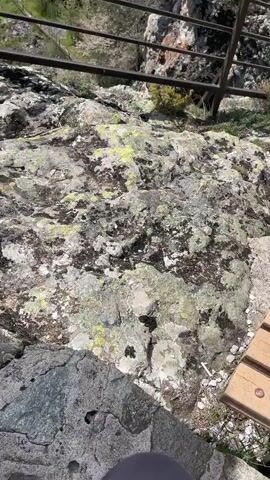 The camera pans up from lichen-covered rocks next to a metal railing. In the distance, a rocky mountaintop can be seen, with a cross on the highest point and a person standing nearby. The view then extends to show a vista of green rolling hills under a sky with fluffy white clouds.
