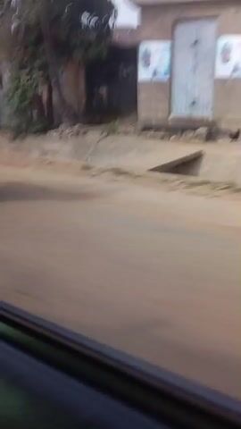 A handheld video shows a blurry street scene, possibly in a developing country. The first shot is a side view of two people on a motorcycle, their faces and clothing indistinct due to motion blur. They are riding on a dirt road. The following shots are also from a moving vehicle, showing the passing landscape. We see buildings with corrugated metal roofs, some appearing dilapidated. There are also people walking in the distance, their features also blurred. A sign with white lettering on a blue background is visible at one point, but the text is illegible. The overall impression is a snapshot of everyday life in a community, captured in a casual and unposed manner.