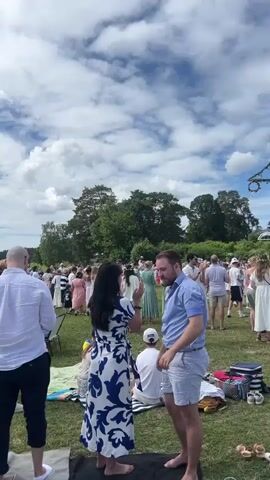 The video captures a large outdoor gathering on a sunny day with scattered clouds. A tall, decorated pole, likely a maypole, stands prominently in the background, adorned with greenery and ribbons. People of various ages are dressed in light-colored clothing, with many wearing white dresses, suggesting a festive occasion, possibly a summer solstice celebration or a traditional Swedish Midsummer event. The attendees are spread across a grassy field, some standing and conversing, while others are seated on blankets or the ground.

In the foreground, several individuals are visible. On the left, a woman with red hair in a yellow floral dress is looking away from the camera. Next to her, a man in a white short-sleeved shirt and dark pants faces away. In the center, a woman in a striking blue and white patterned dress is interacting with a man in a light blue shirt and white shorts. Further back, a group of children are playing.

The overall atmosphere appears relaxed and joyful, with a sense of community and celebration. The lush green trees and the open sky indicate a pleasant summer day. The presence of the maypole strongly points towards a Midsummer celebration, a significant cultural event in Sweden. There is no visible text in the video. The scene unfolds in a spacious, outdoor setting near what appears to be a body of water, with hints of houses in the distance. The time of day seems to be late afternoon or early evening, given the quality of light.