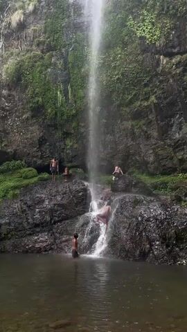 The video shows a tall waterfall cascading into a pool of water surrounded by dark, mossy rocks. Five people are visible, all partially or fully undressed. One person stands waist-deep in the pool, facing the waterfall. Three others stand on the rocks near the waterfall, and one person appears to be climbing on the rocks closer to the falling water. The background consists of a steep, rocky cliff covered in lush green vegetation.