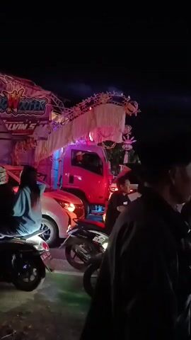 This is a vertical, slightly shaky nighttime video. The scene appears to be a street at night, possibly during a parade or event. In the foreground, several people are visible. On the right, a man wearing a dark jacket and a black cap is in profile, looking towards the left. Next to him, to the left, is another man in a dark jacket, also in profile. Further to the left, a young woman in a light blue jacket is seated on a motorcycle. Two other motorcycles are parked behind her. The main focus of the video is a large, elaborately decorated truck. The truck is red and has a large dragon-like decoration made of white fabric and illuminated with colorful lights that change from pink to green and blue. The truck also has banners and lettering that are partially obscured by the decoration. A white car is positioned in front of the truck, appearing to be involved in an accident or to be part of the procession. Some individuals are standing on the back of the truck, interacting with the decoration. The overall atmosphere is festive due to the lights and decorations, but the presence of the car in front of the truck suggests a potential disruption or incident.