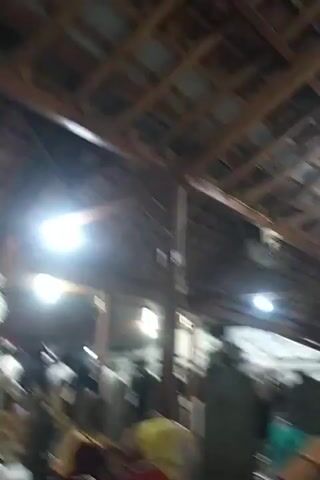 The video shows an indoor scene, likely a place of worship or a gathering space, with a wooden roof structure supported by visible beams. Several bright lights illuminate the area, creating some glare. A number of people are present, appearing to be engaged in various activities. Some individuals are standing, while others are seated. In the foreground, a person is seen from the back, wearing a yellow and red garment, with their head bowed. Further into the scene, there are several vertical stone-like objects, possibly grave markers or decorative pillars, arranged in rows. People are moving around these objects and in the background, suggesting a communal or reverent atmosphere. Some individuals are dressed in light-colored clothing, while others wear darker attire. A ceiling fan is visible in the upper right corner of the frame. The overall impression is of a busy, possibly sacred, indoor environment with a moderate gathering of people.