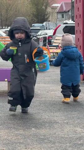 Two small children are at a playground. One child in a dark gray snowsuit with the word "KIDS" on the chest is carrying a small green and yellow cup and a blue bucket full of sand. The child is walking towards the camera and speaks with their mouth open. Another child in a blue coat and gray beanie is walking away from the camera, carrying an orange bucket. The playground has a green and purple slide and a red metal structure in the background. Cars and a building are visible in the background. The ground is gray and appears to be made of rubber.