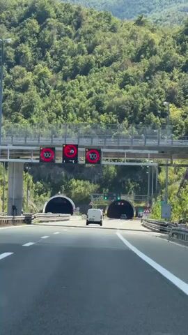 The video shows a view from a vehicle driving on a highway towards two tunnel entrances. Overhead signs display "100" in red circles, indicating a speed limit of 100. One sign also shows a symbol for trucks. There are traffic lights above the tunnel entrances, with green lights illuminated. A white van is visible driving towards the right tunnel entrance. The surrounding environment is mountainous and covered with dense green trees.