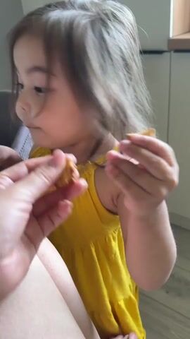 A close-up video shows an adult's hand offering a small, brown, textured snack to a young girl with light brown hair, wearing a yellow sleeveless dress. The girl takes the snack with her right hand and puts it in her mouth at 0:01. She then gently takes the adult's index finger and thumb with her right hand at 0:03 and brings it to her mouth, kissing it at 0:04. The girl then releases the adult's hand and smiles. The background shows light-colored flooring and a white cabinet or wall.
