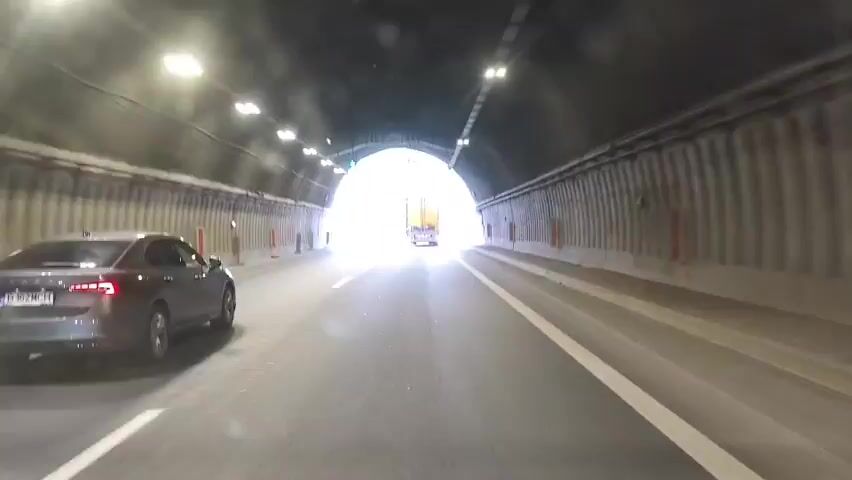 This media file shows a first-person perspective of driving through a tunnel and emerging onto a highway in Vrachesh, Bulgaria. The scene transitions from the dark, artificial lighting of the tunnel to the bright, natural light of the outdoors.

Inside the tunnel, the road surface is grey asphalt with clear lane markings. The tunnel walls are lined with vertical ribbed panels and interspersed with orange and red vertical markers. Lights are visible along the ceiling of the tunnel, and at the tunnel exit, traffic lights with green signals are visible. A silver sedan is driving in front of the camera vehicle, and a white truck is visible further ahead towards the exit.

Upon exiting the tunnel, the road becomes a multi-lane highway with a central reservation. The highway is flanked by a metal guardrail on the right and a grassy embankment with trees and vegetation on the left. The terrain suggests a mountainous or hilly area, with lush green forests covering the slopes. Several traffic signs are visible, including diamond-shaped signs with red borders and white symbols, and a circular white sign with a red border and a diagonal bar. A yellow truck is driving ahead on the highway, and the silver sedan from the tunnel is still visible in front. The sky is blue with scattered white clouds, indicating fair weather. The time of day appears to be daytime, likely late morning or early afternoon given the brightness.

The license plate of the silver sedan is partially visible and appears to read "B 162 MCH". The overall impression is a smooth transition from an enclosed environment to an open, scenic road, suggesting a typical driving experience on a highway bypass. No people are clearly identifiable, but the activity is clearly vehicular travel.