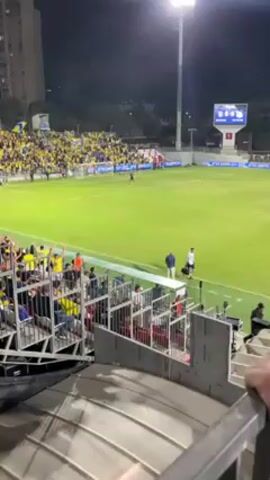The video shows a wide, slightly angled view of a soccer game in a stadium. The perspective is from an elevated position, likely in the stands, looking out over the field. The stadium is well-lit by overhead lights, and the green field stretches out in front. On the left side of the frame, the stands are packed with fans wearing yellow clothing, creating a vibrant block of color. Many fans have their arms raised in the air, suggesting excitement or cheering. In the center of the frame, the field is visible with players in action. A large scoreboard is visible in the background, showing game information. In the immediate foreground, a gray metal railing is present. A hand is seen briefly gripping the rail, suggesting the person filming is holding onto it. The overall environment is energetic, with the visible crowd and the ongoing game creating a sense of lively activity.
