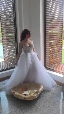 A woman in a white wedding gown with a strapless, beaded top and a full tulle skirt stands between two large windows in a room with white walls and a light-colored floor. Her reddish-brown hair is styled with waves, and she wears a pearl necklace and earrings. She looks to her right, away from the camera. In front of her, on the floor, is a large, light-colored, shell-shaped bowl containing a bottle of white wine, a smaller red box, and other items. The windows are covered with horizontal wooden blinds.