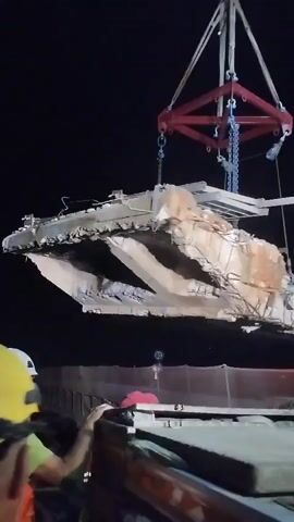 A large, damaged section of concrete, possibly from a bridge or building, is being lifted by a crane at night. The concrete is jagged and broken, with exposed rebar. Below the concrete, a flatbed truck is visible, with people standing on it and nearby. One person is wearing a green shirt and a white hard hat. The scene appears to be a demolition or salvage operation.