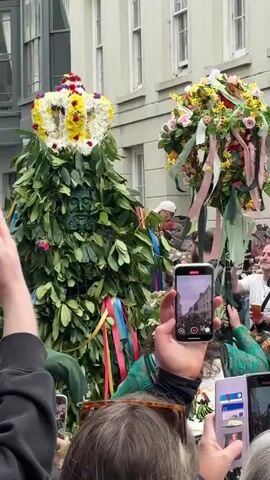 A medium shot shows a street parade or festival with a large crowd of people in the street taking photos with their phones. The focus is on a person dressed as a "Green Man" figure covered in leaves, with a crown of flowers on top and ribbons hanging down. There is another person nearby also covered in flowers. The architecture of the buildings on the street is visible, with a shop sign that reads "Roberts Curios." It appears to be a lively outdoor event with many attendees.