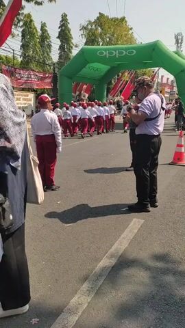 The video shows an outdoor event with several people. In the foreground, a person wearing a light purple shirt, dark pants, and a camouflage hat is standing and looking towards a group of children. The children are wearing matching red and white uniforms and appear to be participating in a parade or a procession. They are walking in formation under a large green inflatable arch that has the "Oppo" logo displayed on it. There are also some traffic cones on the road, suggesting a controlled area for the event. The background shows trees and buildings, indicating an urban or suburban setting. The lighting suggests it is daytime and sunny. The camera pans slightly to the right throughout the video.