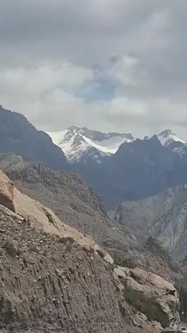 The video shows a view of a mountainous landscape, likely taken from a moving vehicle. The foreground features a rocky hillside with sparse vegetation. In the midground, there are several layers of mountains, some partially obscured by haze. In the background, the mountain peaks are covered in snow. The sky is partly cloudy with patches of blue visible. Later in the video, a power line tower comes into view, positioned on the rocky slope. The overall lighting suggests daytime, but the clouds create a somewhat diffused light. The camera movement is relatively smooth, implying a stable camera platform, possibly inside a car.