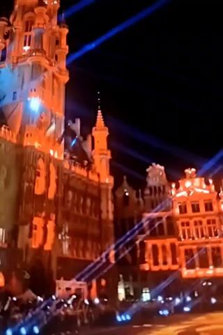The video shows a nighttime view of a city square decorated with lights. Ornate buildings, illuminated in orange and white, surround the square. Blue lights are projected across the buildings and into the dark sky. In the foreground, a large floral carpet made of what appears to be dried flowers is visible, illuminated by orange and red lights. The carpet has a geometric pattern. A crowd of people, silhouetted against the lights, are gathered in the square. The camera pans across the scene, capturing the architectural details of the buildings and the vibrant light display.