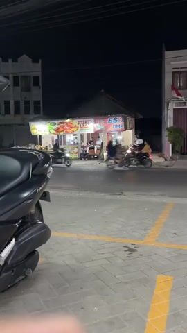 At the beginning of the video, a black scooter is parked in the foreground on the left. The scooter is facing away from the camera. Behind the scooter, a street is visible, with a small food stall on the right side of the street. The food stall is lit up and has people standing around it. There are several motorcycles parked near the food stall. On the left side of the street, there is a building under construction. A few people are visible in the background, walking or standing near the food stall. As the video progresses, a red truck with a green tank drives across the street from right to left. The truck is moving at a moderate speed. The lights of the truck are illuminated. The food stall continues to be illuminated, and people are still visible around it. The black scooter remains parked in the foreground. The street is paved with interlocking bricks, and yellow lines are painted on the ground. The sky is dark, indicating that it is nighttime. The buildings are simple structures, with one appearing to be multi-story. The overall scene depicts a busy street scene at night with a food stall and passing traffic.