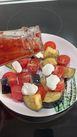 A white plate with a green leaf pattern around the rim is filled with quartered cherry tomatoes, chopped cooked eggplant, and several dollops of white soft cheese. A glass bottle of red sauce is being held over the plate, pouring sauce onto the food. The plate is sitting on a black stovetop, and a white kitchen counter is visible in the background.