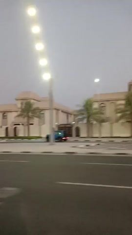 A grainy, low-light video captures a street scene with multiple large, modern houses lining the right side of the road. The houses are predominantly white with some beige accents and feature palm trees in front of them. A light post with multiple bulbs is visible on the left side of the road, illuminating the street. A blue SUV is seen driving on the road, and a white RV is parked in front of one of the houses. The sky is overcast, suggesting either early morning or late evening. The camera is moving, indicating the video was likely filmed from a vehicle.
