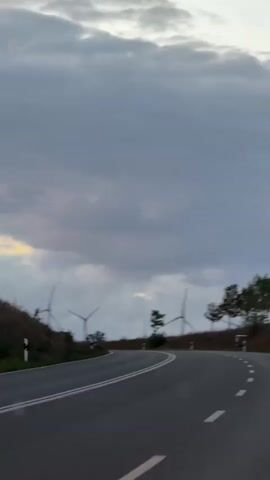 A car is driving on a winding asphalt road with white dashed lines. The sky is cloudy with some light breaking through. There are several wind turbines visible on a hill to the left and in the distance. The landscape is hilly and covered with grass and trees.