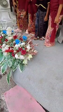 The video opens with a close-up of a floral arrangement of white, red, and blue flowers, with lush green leaves. The arrangement is placed on a grey surface, possibly concrete or stone, with a pink object partially visible in the foreground. As the camera pans up and moves to the right, a group of people are seen standing. They are dressed in traditional attire. Several women are wearing headscarves and patterned sarongs. Among them, there are children also in traditional clothing, including maroon tops and patterned sarongs. A man is visible wearing a dark, ornate outfit, possibly a suit or ceremonial attire, and a woman in the background is wearing a gold headdress, suggesting a wedding or formal cultural event. The environment appears to be indoors, with a corrugated metal ceiling visible. The lighting is bright, suggesting daylight or artificial lighting. The people are mostly looking towards the center, where the bride and groom might be. The overall scene depicts a formal gathering, likely a celebration or ceremony.