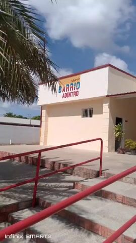 The video shows an exterior shot of a building with a sign that reads "Misión BARRIO ADENTRO" and "CENTRO MEDICO DE ALTA TECNOLOGIA Dr. DAVID QUENDSECHADZE". The building is painted in light colors with red trim. There are steps leading up to the entrance, with red handrails.  There are potted plants near the entrance. People are sitting outside the building. In the background, there is a palm tree and a view of distant mountains. The sky is blue with scattered clouds. The video is slightly shaky, suggesting it was taken while walking. The bottom of the screen displays "Infinix SMART 8."