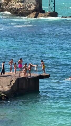 A group of young people are gathered on a rocky ledge overlooking the ocean. One person, wearing yellow shorts, is in mid-air, having just jumped off the ledge into the water. Another person is already swimming in the water. The water is a clear turquoise color, and there are waves crashing against the rocks in the background. The ledge has a metal railing. Some people are standing and watching, while others appear to be waiting their turn to jump. The sky is clear and the sun is shining.