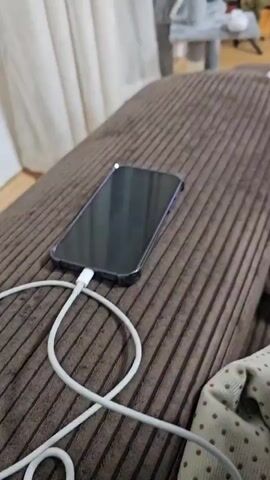 The video captures a close-up of a dark-colored smartphone lying on a brown, ribbed fabric surface, likely a sofa or bed. The phone is encased in a dark protective cover and is connected to a white charging cable. In the background, a light-colored curtain and some blurry furniture, possibly a cat tree, are visible. The camera then quickly zooms in on the smartphone's screen, which is black and off. A faint purple reflection is briefly seen on the phone's edge as the camera approaches. The screen surface shows minor smudges or dust particles. No people are clearly visible, and no text is discernible on the phone or in the immediate surroundings. The scene is set indoors.