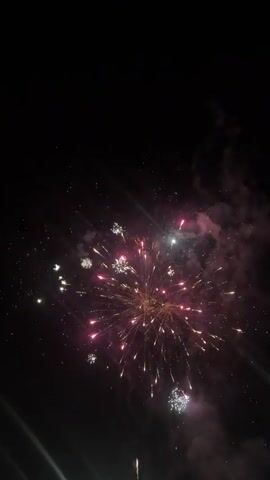 The vertical video displays fireworks exploding against a dark night sky. The primary explosions are a cluster of white and silver starbursts, filling the upper portion of the frame with bright, sparkling lights and trails of smoke. Interspersed with these white bursts, there is one instance of a large, bright yellow-gold firework explosion that creates a central burst of light and sparks. The explosions occur in quick succession, with lingering smoke partially obscuring the view between bursts. A few ascending fireworks and distant sparks are briefly visible in the darker moments.
