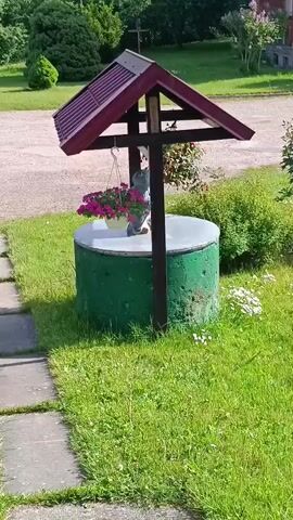 A video shows a decorative well in a garden. The well has a green concrete base and a red-roofed wooden structure above. A pot of pink flowers hangs from the roof. A stone path leads to the well, which is surrounded by green grass and bushes. The sun is shining.