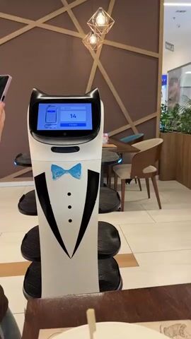 A service robot, designed to resemble a cat wearing a tuxedo, stands in what appears to be a restaurant or cafe. A person's hand, holding a smartphone, is visible on the left side of the frame, interacting with the robot. The robot has a screen displaying a numerical value of "14" and a "Finish" button. The robot has multiple trays for carrying items. In the background, there are tables and chairs, and decorative lighting fixtures are suspended from the ceiling. The environment suggests an indoor setting with modern decor.