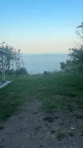 This is a video file showing a scenic view of the sea from a grassy hilltop. The scene is tranquil and serene.

The primary subjects are the natural elements: the vast expanse of the sea, a clear blue sky that transitions into a lighter hue near the horizon, and a grassy area in the foreground with some scattered vegetation and bare branches of trees.

There is a single, notable object in the middle ground: a rusty, reddish-brown barrel or container standing upright on the grassy slope overlooking the sea. It appears to be the only man-made object prominently featured in the natural landscape.

The activity taking place is minimal, suggesting a moment of stillness and observation. The camera is stationary, capturing a static view of the seascape. A tiny, dark speck that might be a bird is visible in the sky.

The time of day appears to be either early morning or late evening, judging by the soft, diffused light and the pale colors of the sky. The weather seems calm and clear, with the sea surface showing gentle ripples. The location cues, combined with the provided context of Osenovo, Bulgaria, suggest a coastal setting in this region. The overall emotion conveyed by the scene is one of peace and quiet contemplation, emphasizing the beauty of nature.

No visible text is present in the video.