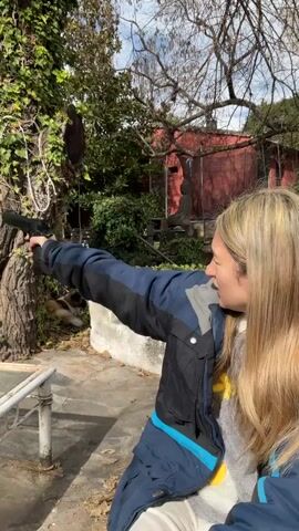 A woman with blonde hair is sitting outdoors, holding a gun. She is wearing a blue and black jacket over a gray sweater and white pants. She is aiming the gun at a tree with green leaves. In the background, there is a red building with a statue in front of it. The ground is covered with fallen leaves.