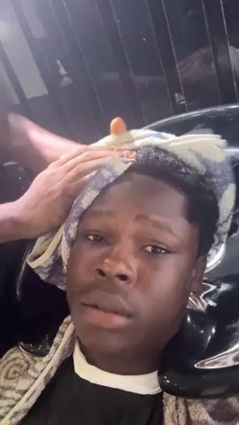 A close-up shot shows a young Black person with dark hair and dark eyes sitting in a salon chair, leaning back. Their head is resting against a black salon sink, and a white towel with blue stripes is draped around their neck and over their shoulders. Another towel, also white with blue stripes, is being used by an unseen person to wipe the young person's forehead and temples. The young person's face is illuminated, and they appear to be looking upwards. In the background, vertical black panels are visible, reflecting light. The overall scene suggests a hair washing or treatment service.
