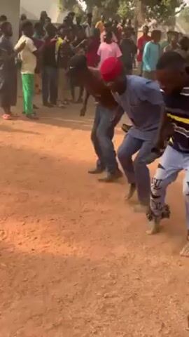 A medium shot of a group of men dancing in a dusty area outside.  The men move in a coordinated line, bent at the waist, kicking up dust as they move.  The ground is red dirt.  Most of the men wear casual clothing, with one sporting a red fez.  They are surrounded by a crowd of onlookers.  In the background, a white building is visible.  The camera shakes slightly, indicating it’s handheld.  The overall scene appears festive and energetic.