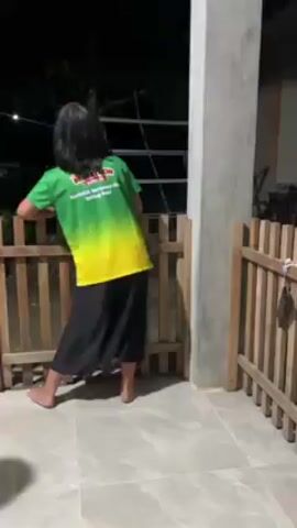 The video shows two young children standing on a porch in front of a wooden fence at night. The older child is wearing a green and yellow shirt and black pants and is facing away from the camera, leaning on the fence. The younger child is wearing a white shirt and green patterned pants and is walking around. The porch is made of light-colored tile and has a concrete pillar on the right side. A tree and some houses can be seen in the background. The lighting is dim, with a light source above and to the left of the children. There is audio playing.