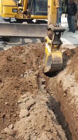 An excavator is digging a trench on a street. The excavator's yellow bucket is digging into the brown dirt, throwing soil to the side. The trench is about waist-deep and has steep sides. The excavator is yellow and has a black blade at the front. A person in a blue shirt and jeans is standing on the sidewalk behind the excavator, watching. There are some buildings in the background.
